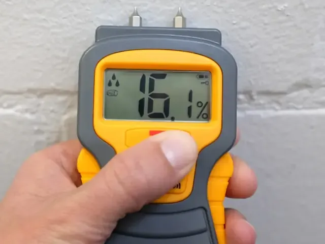A handheld moisture meter measures moisture against a textured interior wall